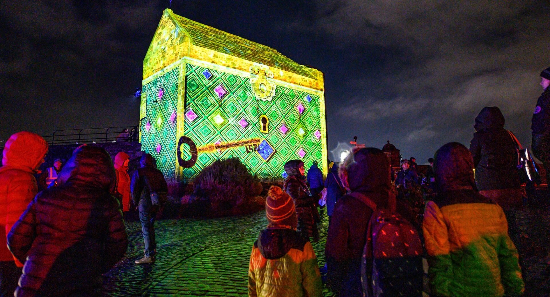 Part of Edinburgh Castle lit up yellow and green like a treasure chest, with a large key on the front. There are crowds in the foreground