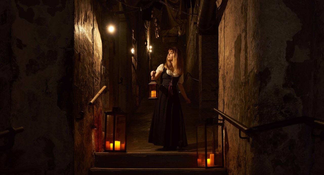 A woman carrying a lantern walks down the steps of a dark, candle-lit close