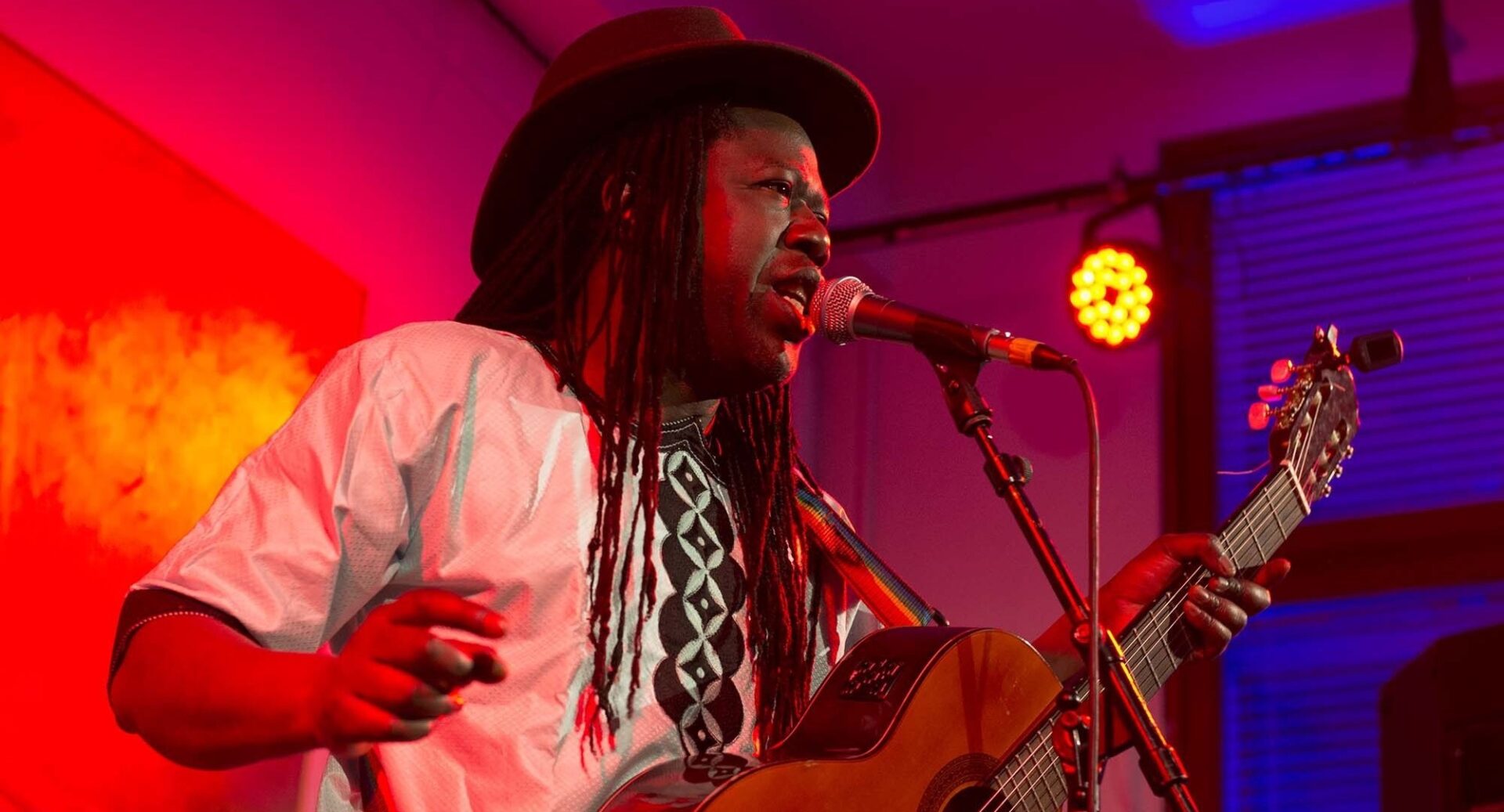 A singer wearing a brown trilby, white shirt and with long dreadlocks sings into a microphone as he holds his acoustic guitar