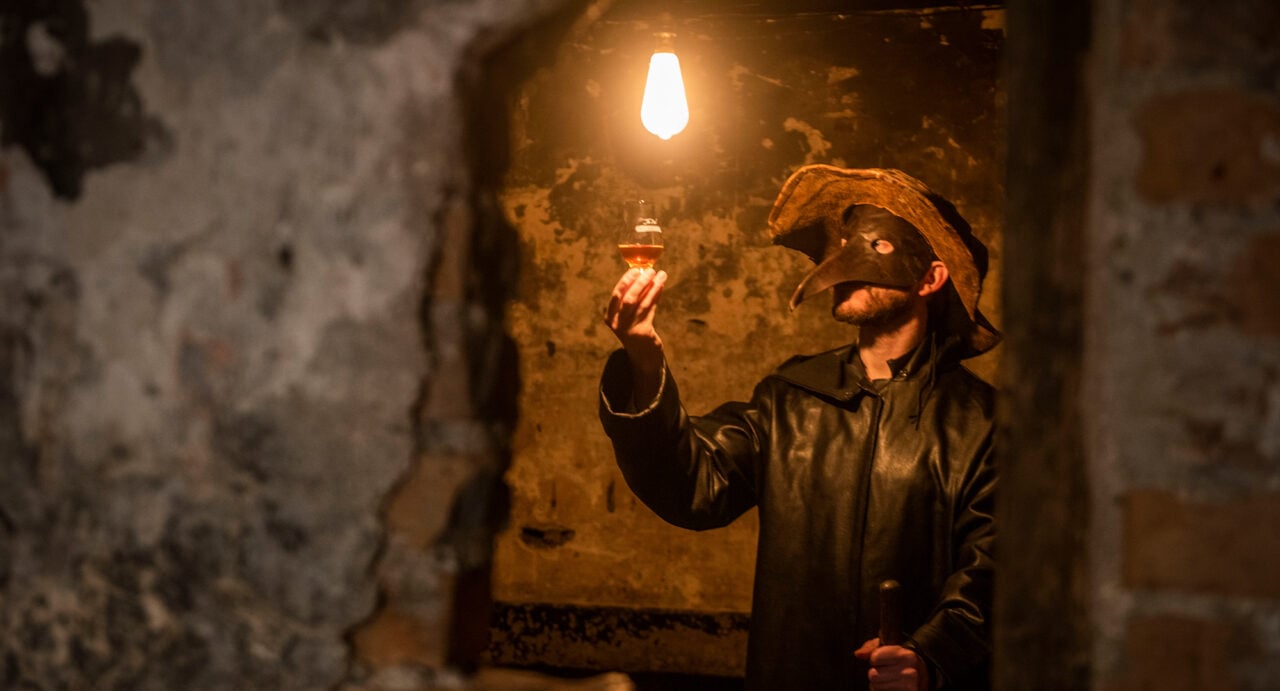 A performer wearing a leather coat, hat and plague mask holds up a glass of whisky to the light