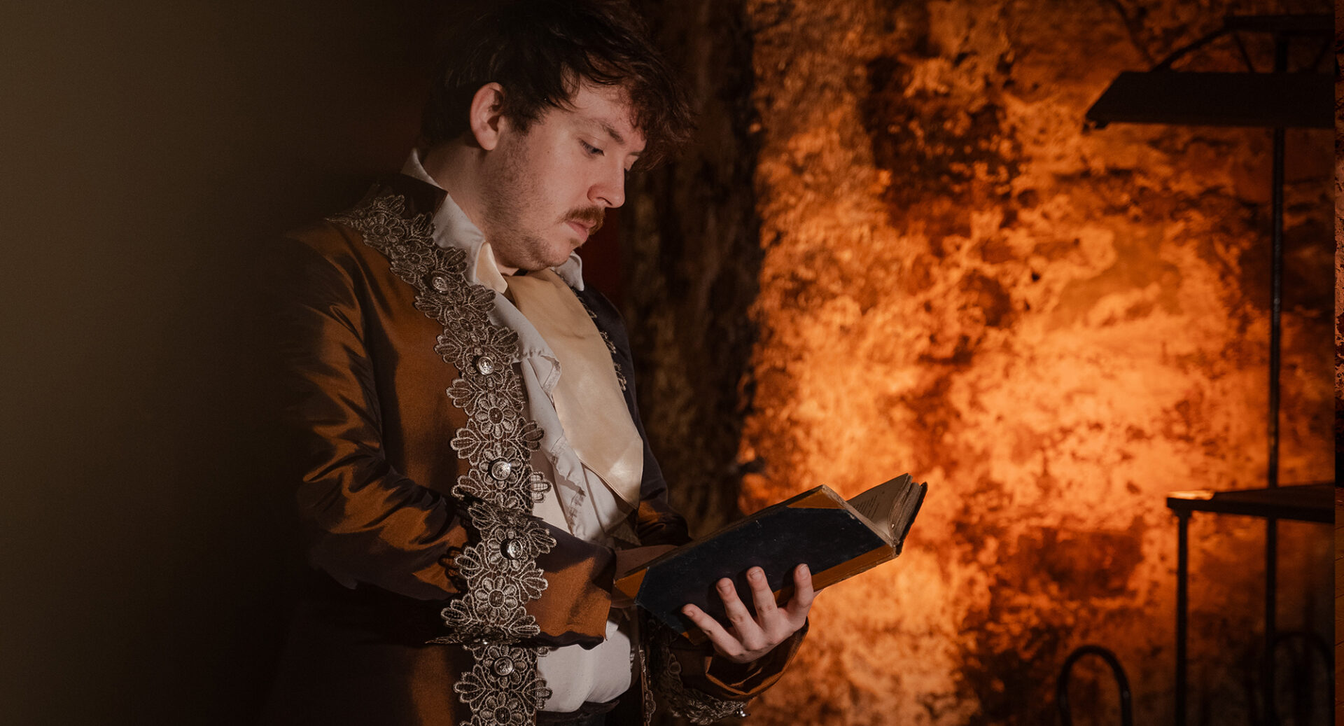 A performer in an ornate bronze jacket with a large beige cravat reads from a book of poetry