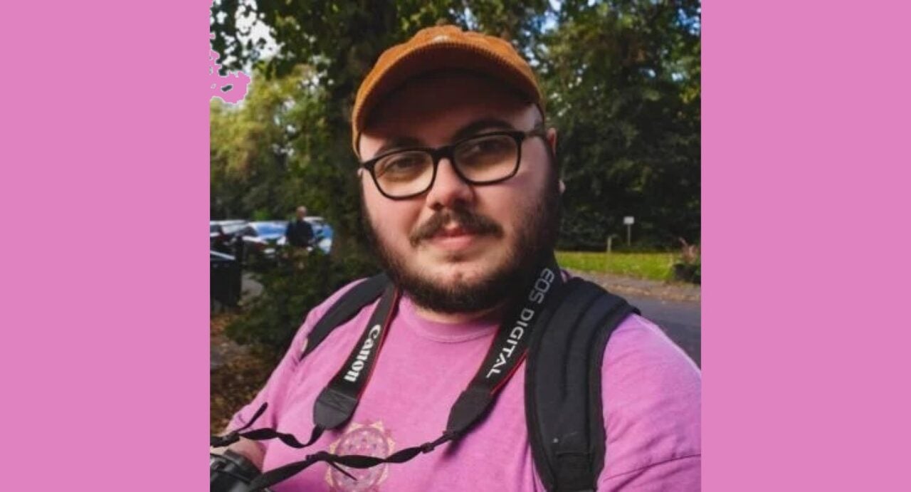 Aaron Sheridan headshot. He wears black rimmed glasses, a pink tshirt, an orange baseball cap, and has a camera strap around his neck