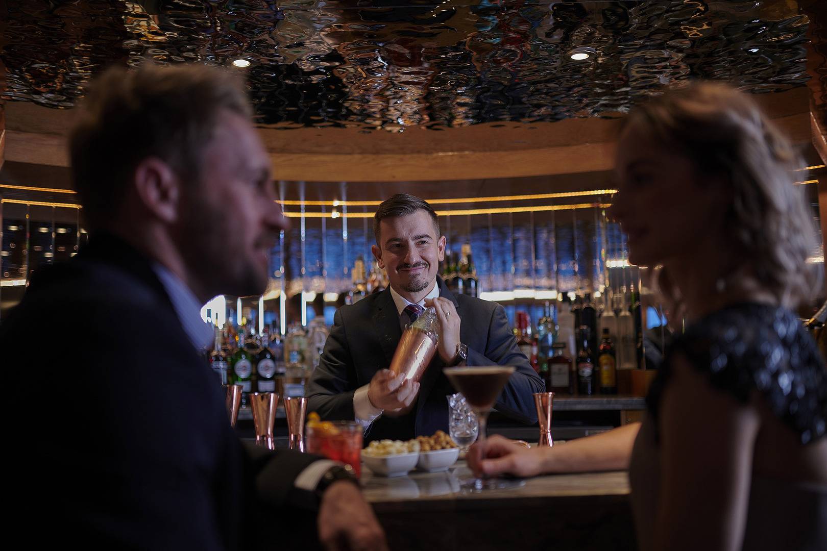 A man and a woman sitting at the bar as a member of the Fingal crew makes a cocktail.