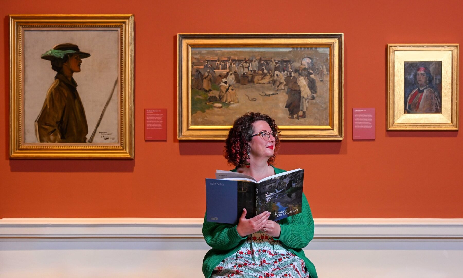 Lady holding a book about Sir John Lavery with three of his paintings displayed behind her