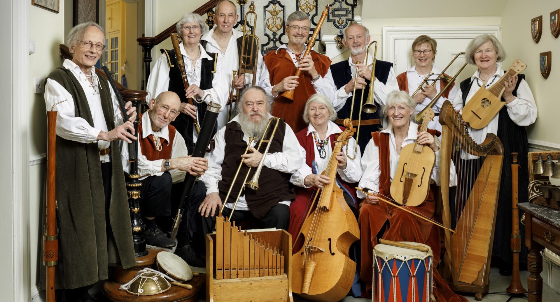 Eleven people in mediaeval-type clothing with a selection of ancient musical instruments including a harp, drum, and wooden and metal string and wind instruments