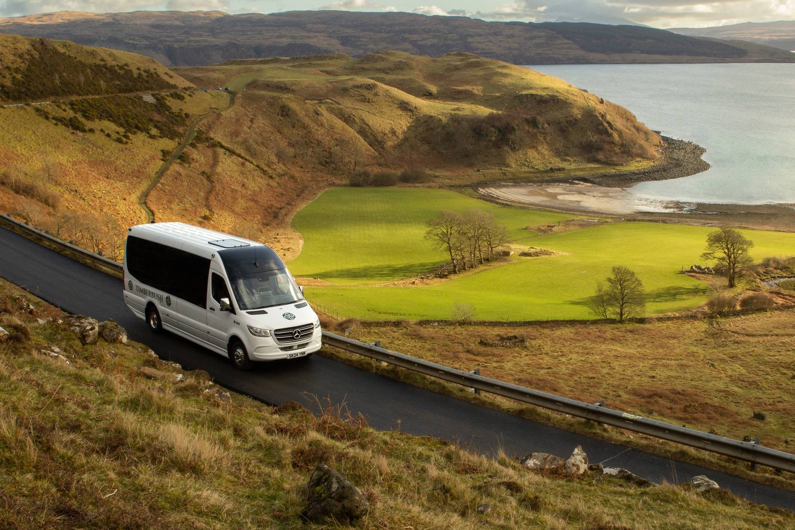 Timberbush coach on single track road in the Highlands of Scotland, with mountains and a loch in the background.