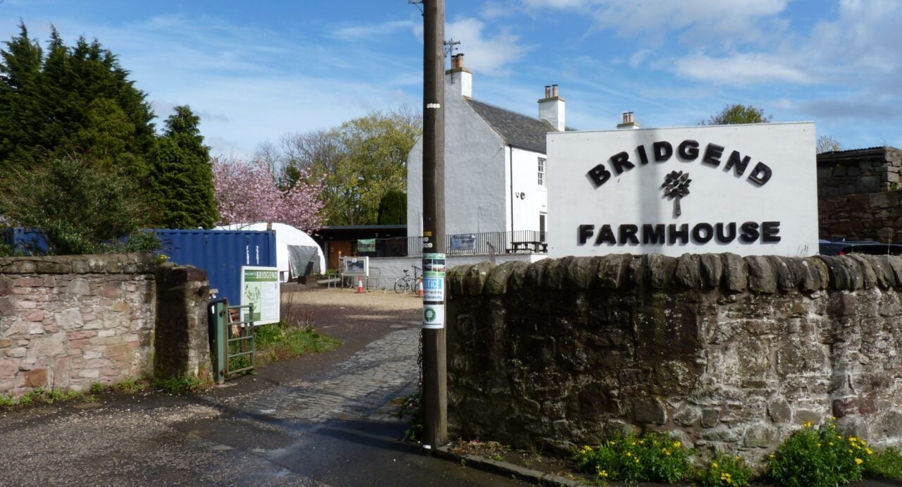 The entrance to Bridgend Farmhouse, with the sign on an old stone wall on the left, and trees beyond the stone wall on the left and beyond. Ahead up the path can be seen a white buildikng surrounded by railings and with outdoor seating to the front