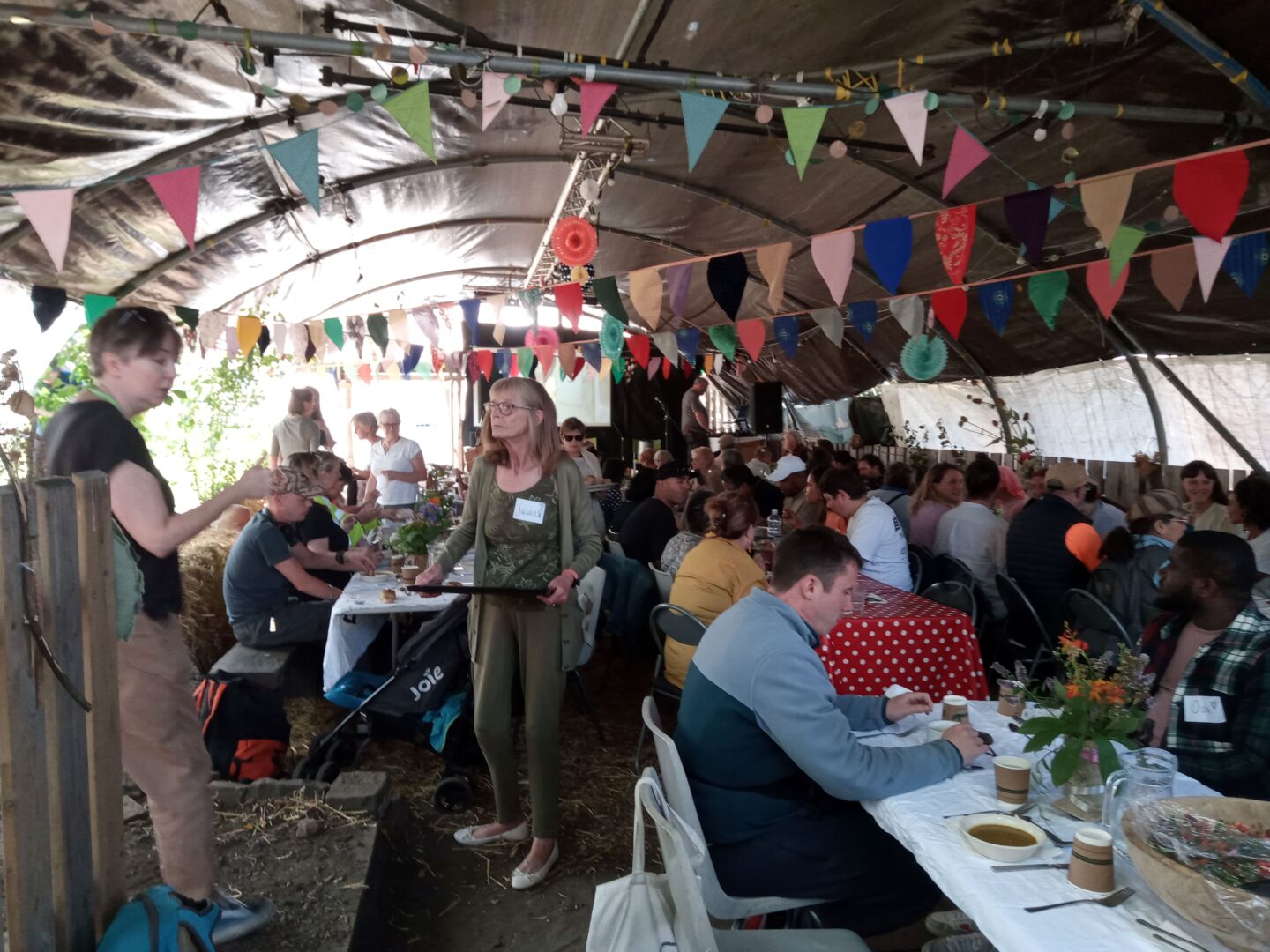 Crowd of people sitting on shared tables sharing a meal. Bunting hangs overhead.