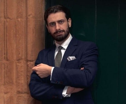 A shot of Dr Sam Gallacher in suit and tie leaning against the entrance of a building