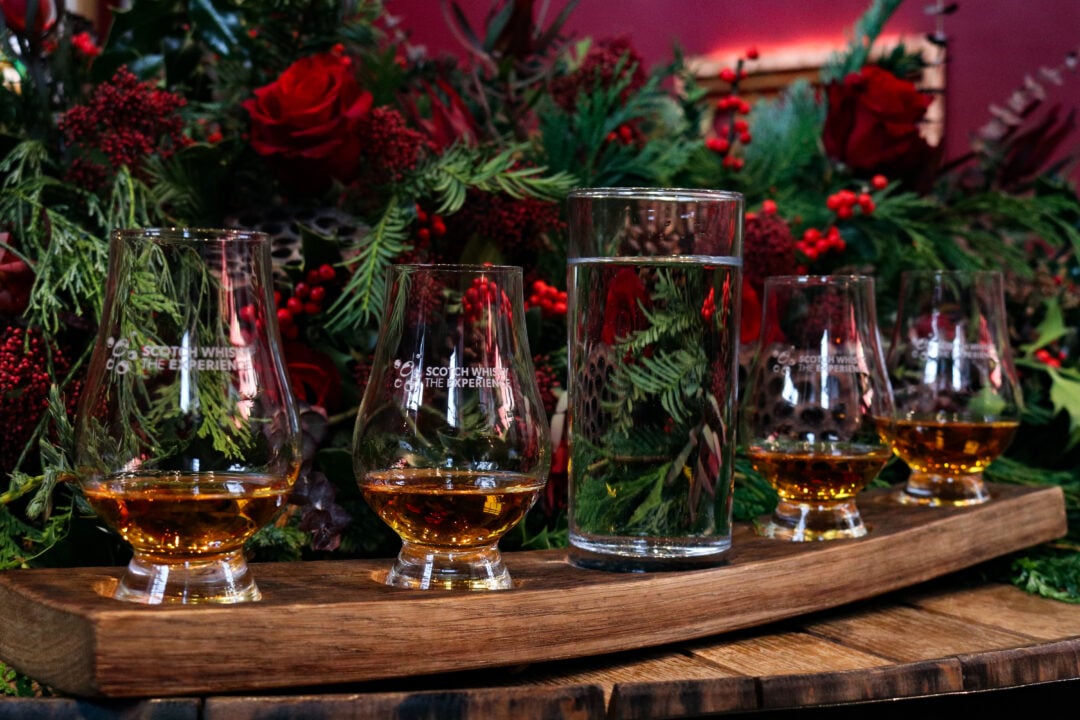 4 glasses, partially filled with whisky, sitting on wooden tray. Christmas garland with berries in background.