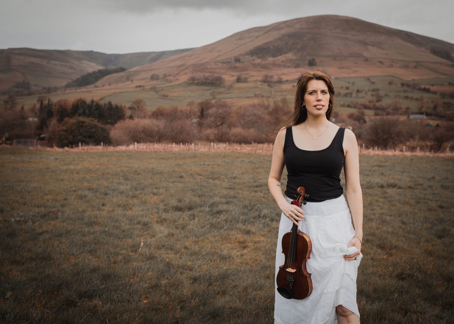 Women wearing black vest and white skirt standing with violin in one hand, against a hilly, mountain background.