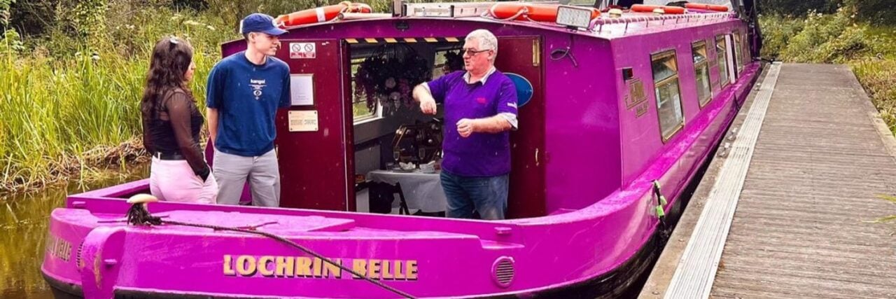 The pink Lochrin Belle canal boat on the canal, with three people standing at the rear