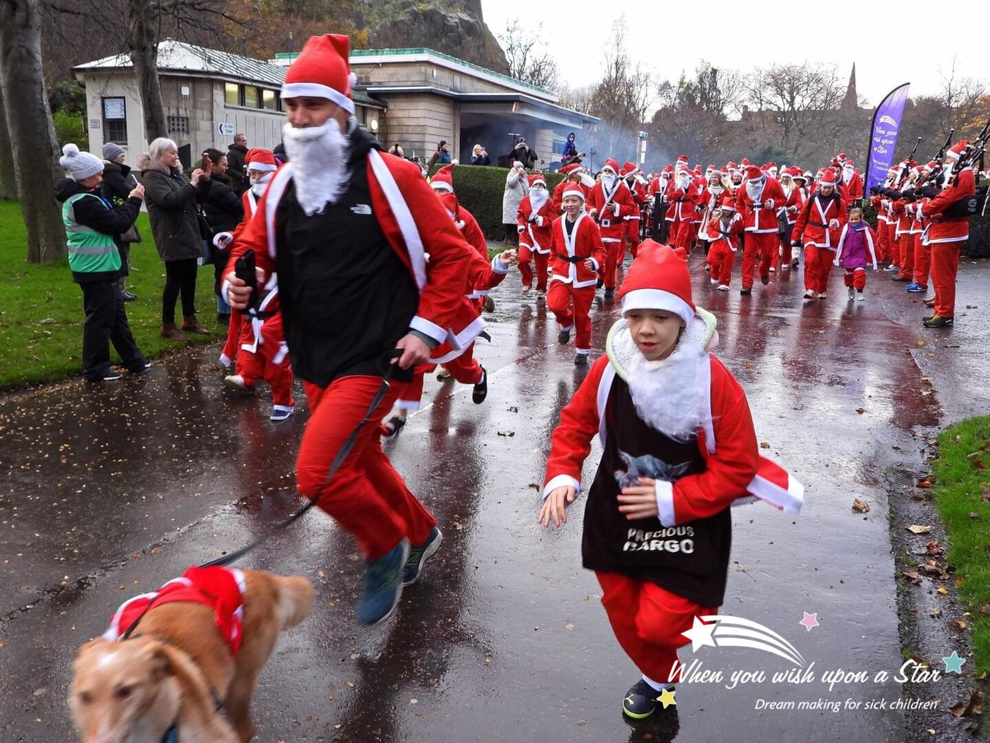 Group of adults and children dressed in Santa outfits running around park.