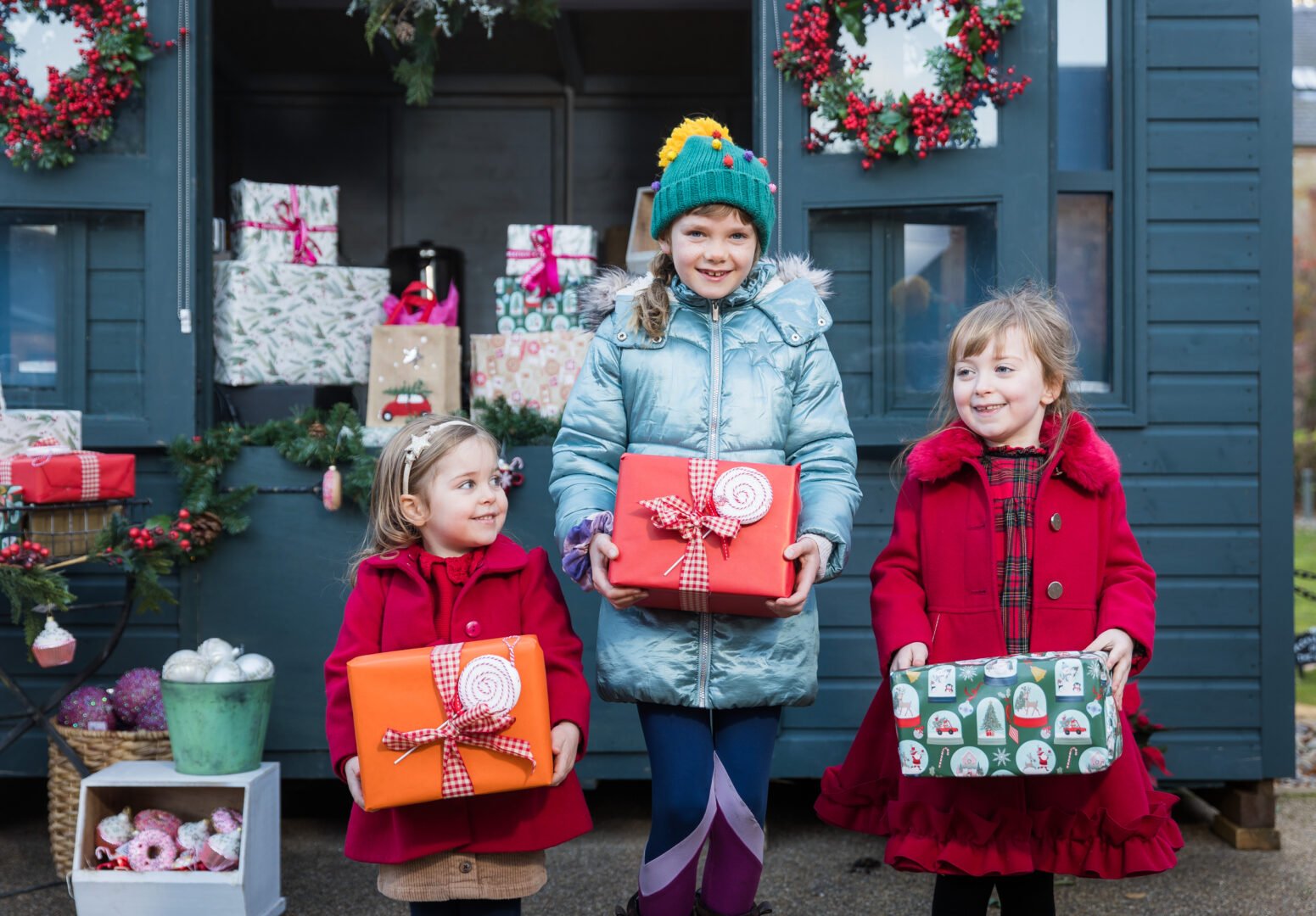 3 young girls, dressed in winter jackets and each holding a wrapped present, standing in front of a shed, with it's windows open and wrapped parcels displayed inside.