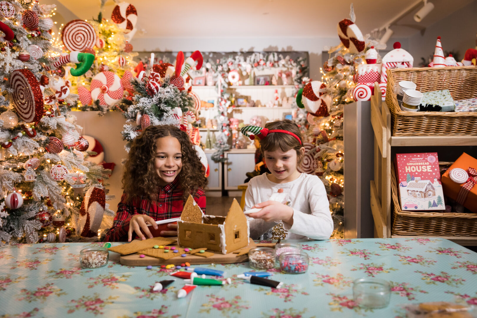 2 young girls sitting at a table, making a gingerbread house. The background is decorated with festive decorations.