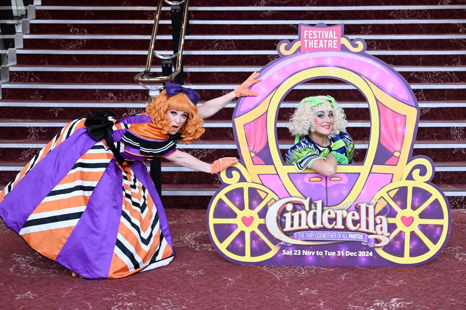 One women pretending to push a cut-out of an elaborately decorated pumpkin coach, inside of which sits another women.