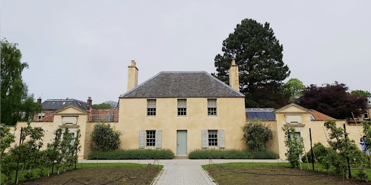 Front-facing image of a pale yellow painted 2 story house, with 2 chimneys and grey tiled roof, set in a paved garden.