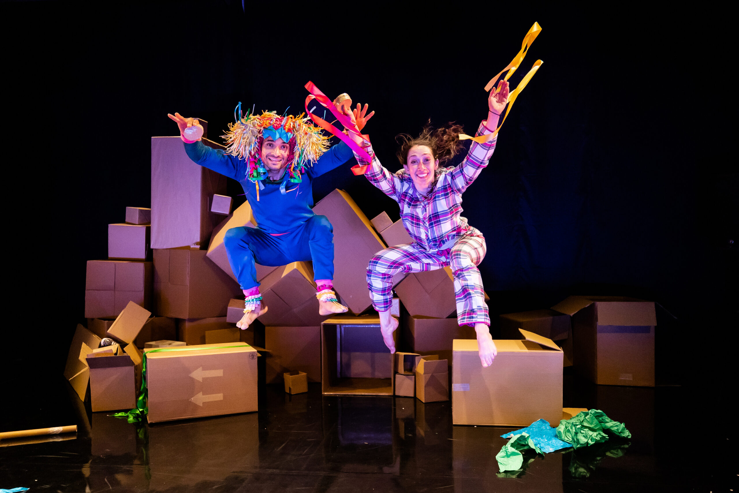 2 people, dressed in pyjamas, jumping in the air. Behind them are a pile of cardboard boxes.