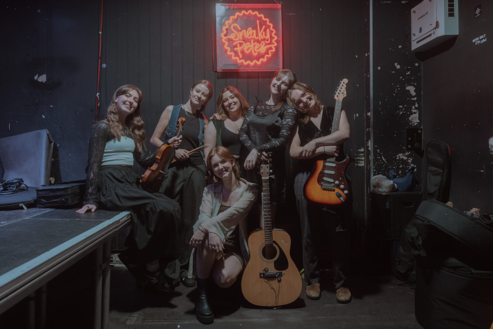6 females standing in a group. There is a guitar on the floor in front of them.