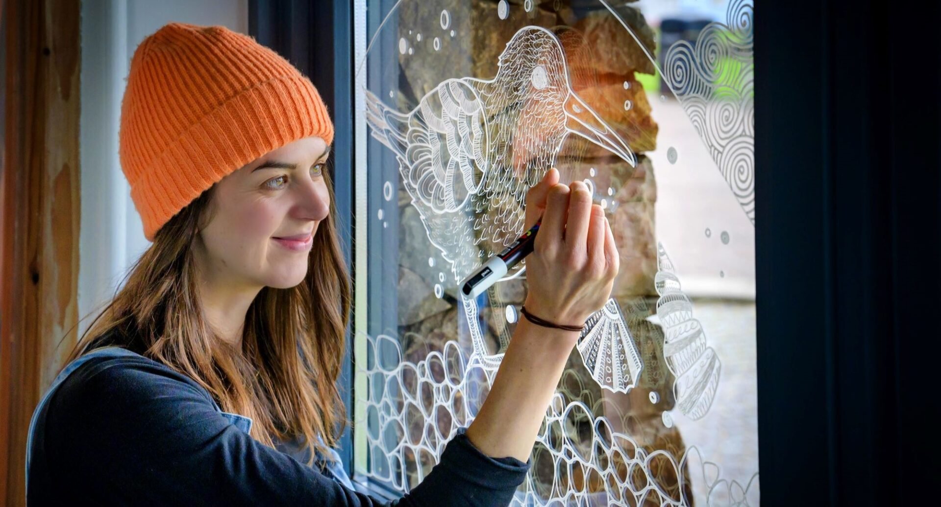 Girl with brown hair and an orange hat drawing with an image of a seabird with white pen onto a clear window pane.