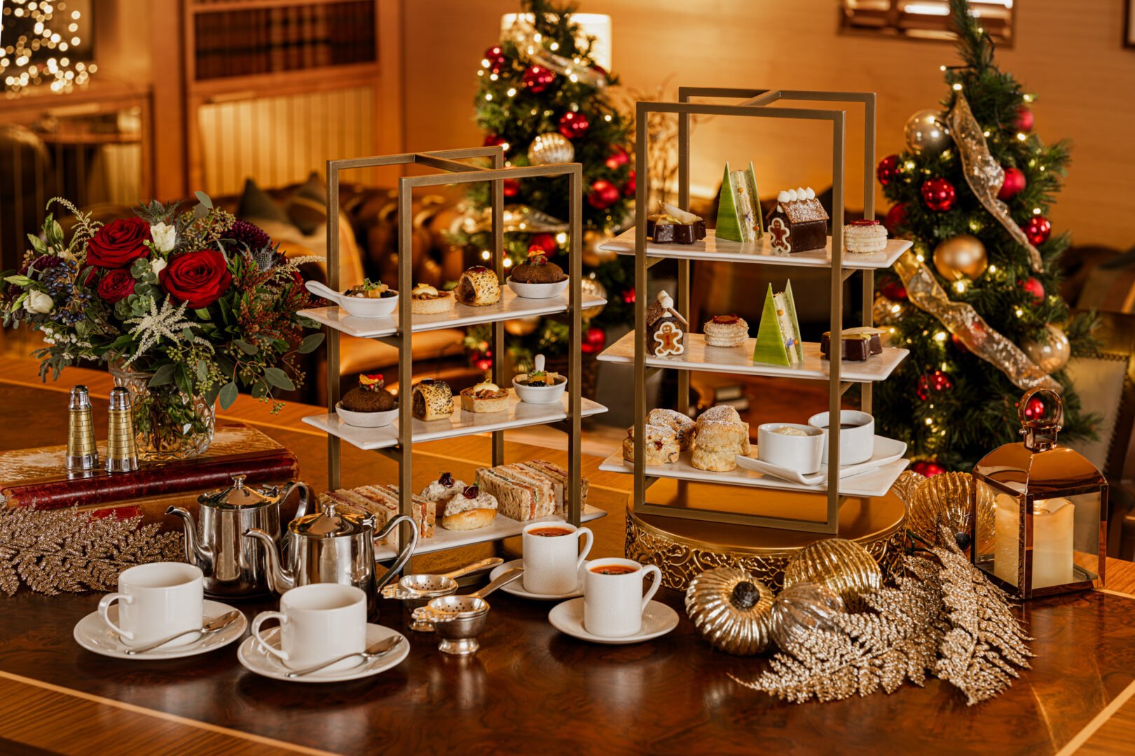 A table set with 2 tiered cake stands, filled with cakes and scones and sandwiches. In the background are 2 decorated Christmas trees.