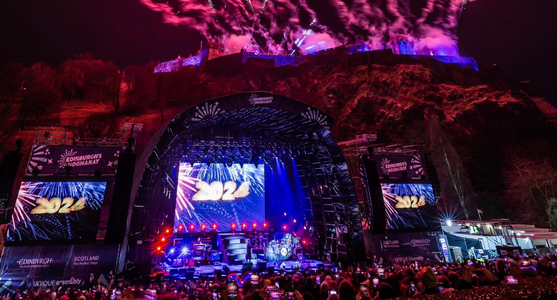 Crowd looking at giant illuminated screens, against backdrop of a castle perched of a clifftop.