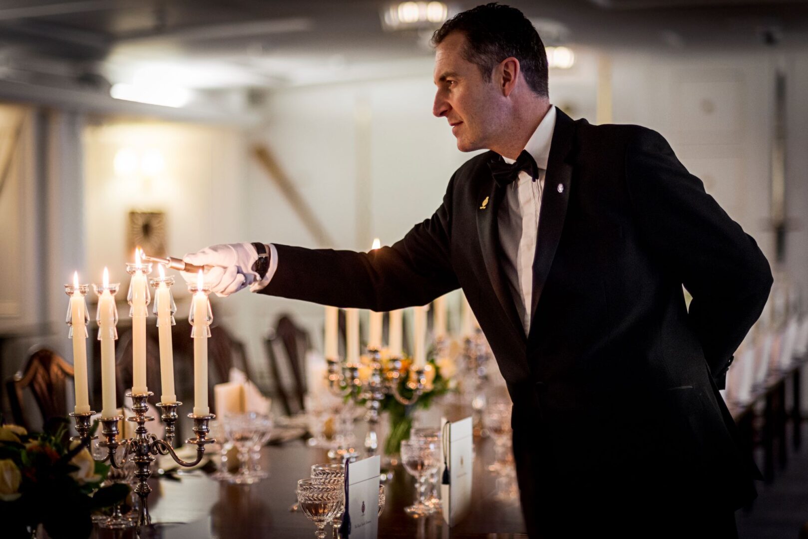 Man in dark suit lighting a candelabra, which is sitting on a decorated dining table.