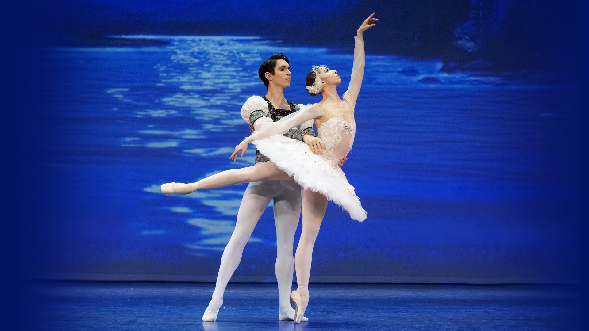 2 dancers on stage, female wears a white tutu and is being supported by male partner as she reaches one arm into the air.