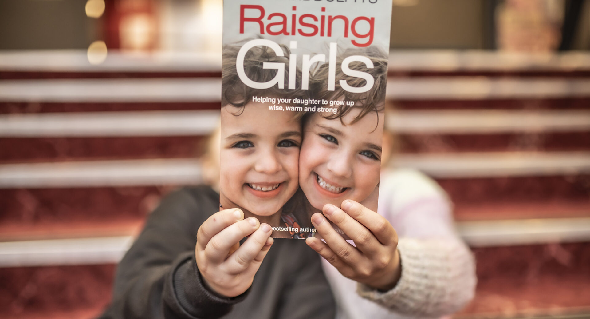 Twho children holding a Raising Girls book in front of their faces. A staircase is behind them