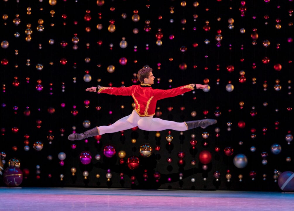 Male dancer in red jacket and white trousers leaping across the stage.