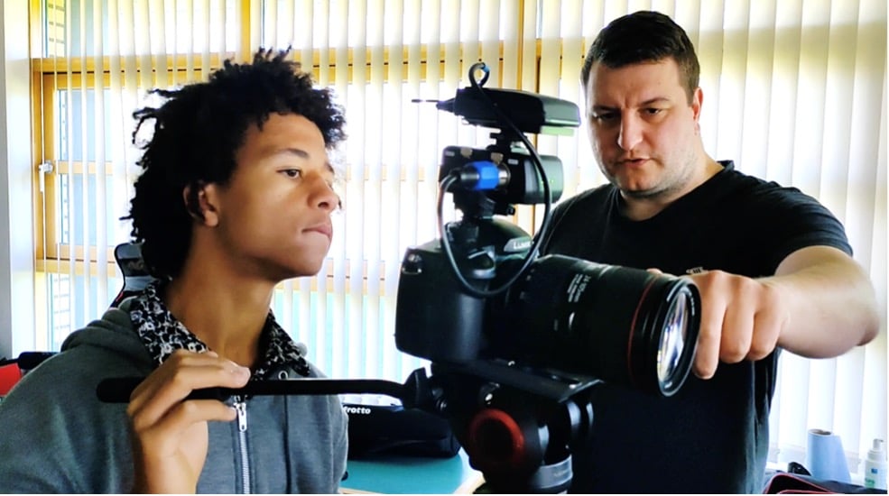 A man in a black tshirt showing a younger man how to operate an SLR camera on a tripod