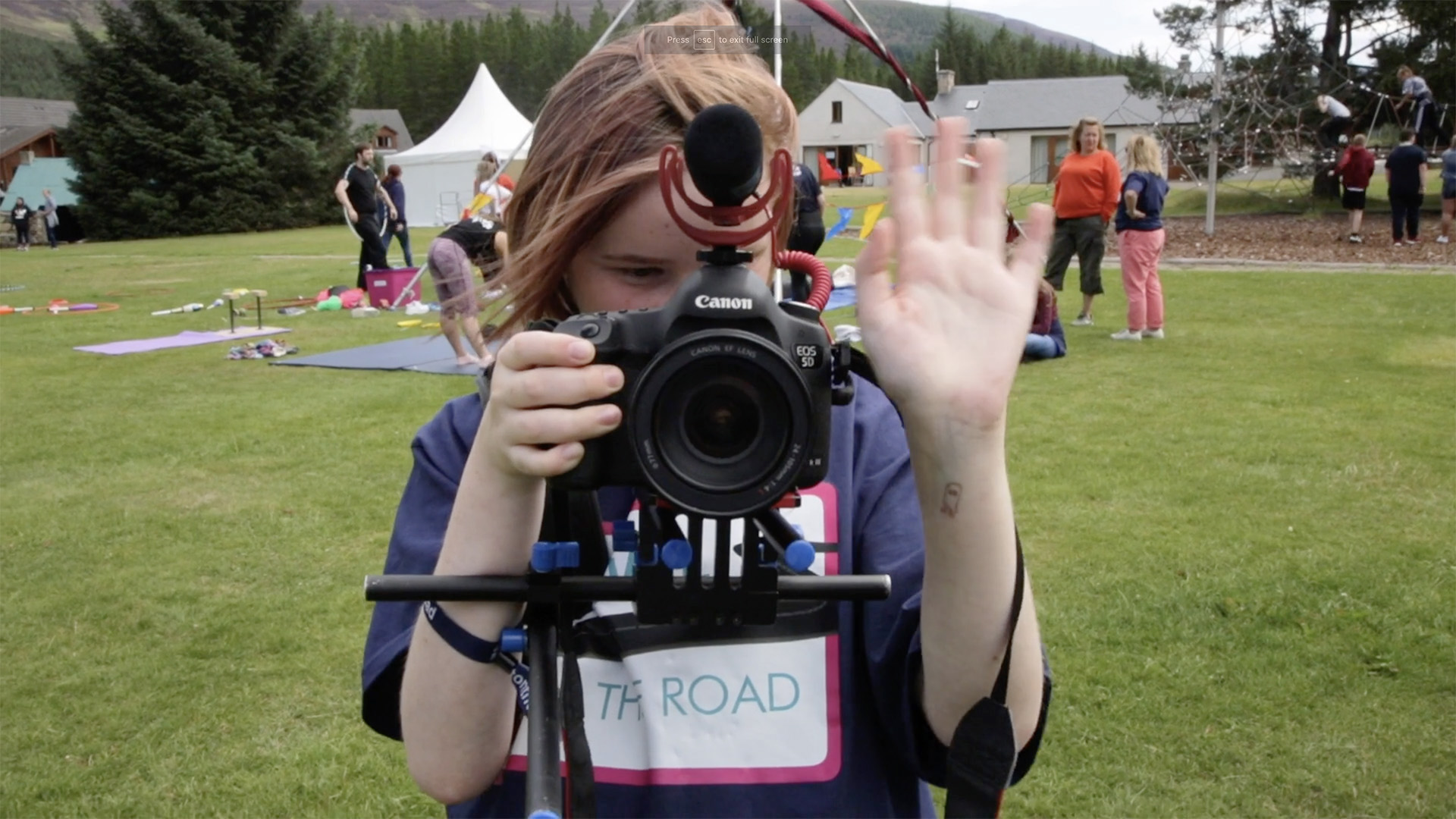 A young girl behind an SLR camera looking through the viewfinder and waving. People , bunting and a marquee are in the background in what looks like a public park