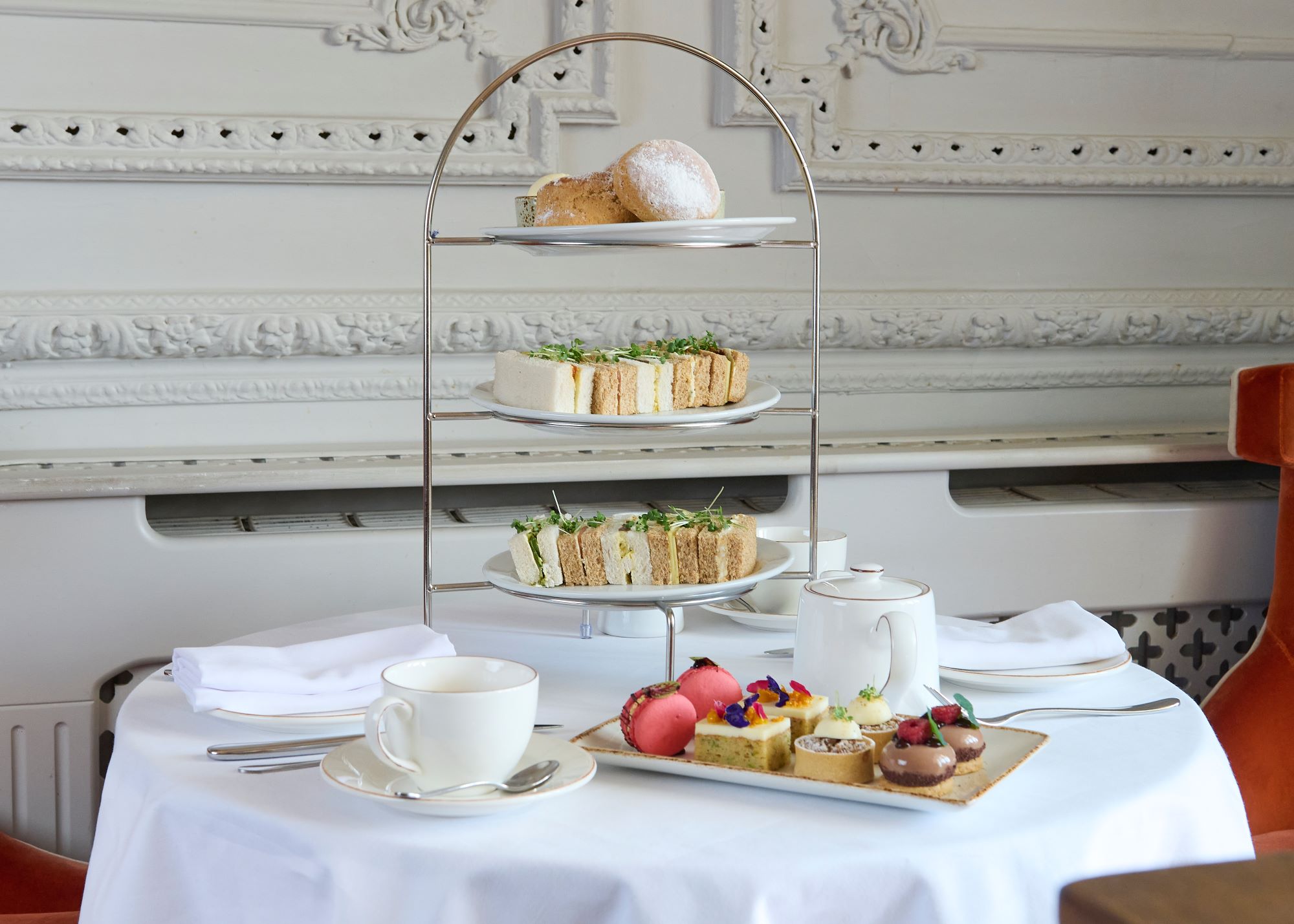 3-tier cake stand filled with scones and sandwiches sitting on a table. The table is also set with a white teapot, 2 cups and saucers, white napkins and a plate of small cakes.