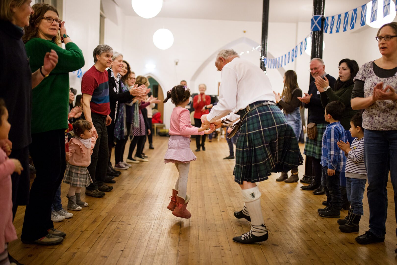 A man in a kilt holding a little girl's hands as they dance between two rows of clapping onlookers. Saltire bunting is hanging from pillars.