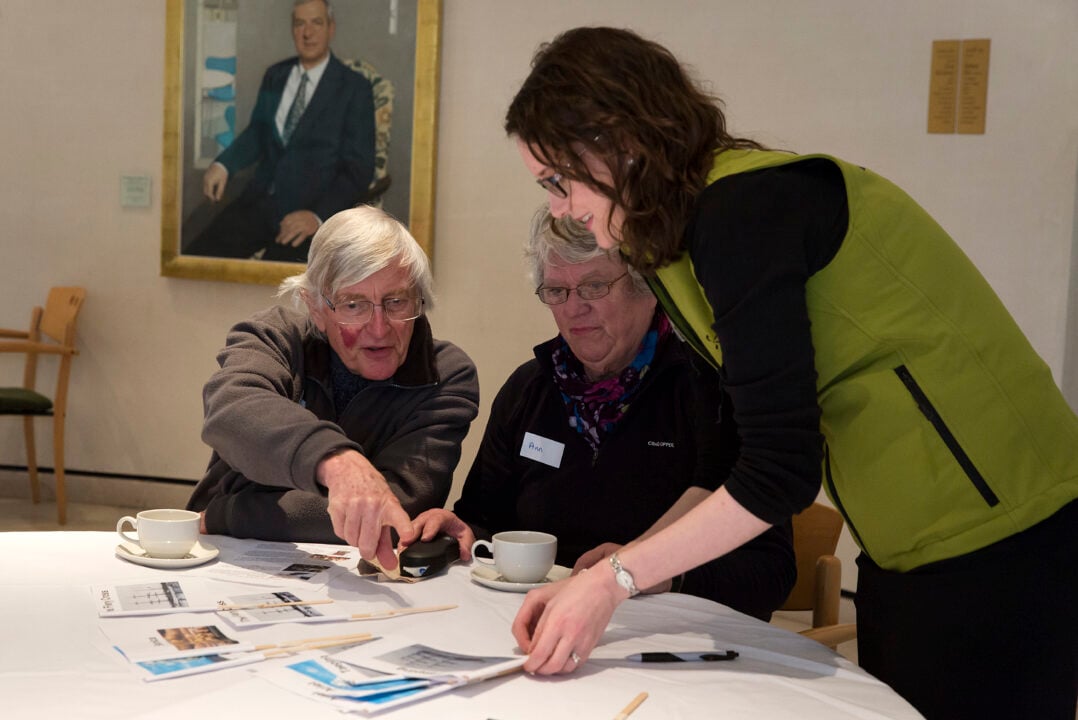 A couple sat at a table looking at pieces of paper, while a woman in an olive green bodywarmer stands beside them.