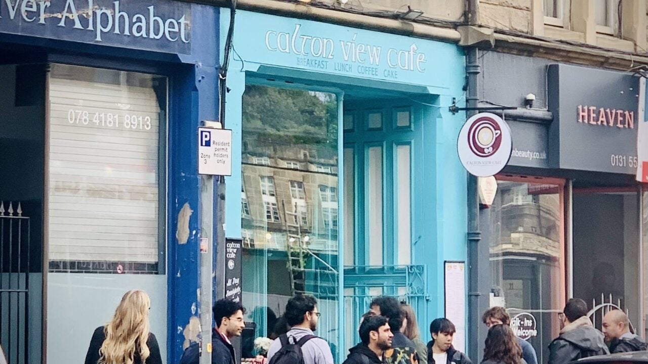 A vibrant cyan cafe with a bustling crowd gathered outside, enjoying the lively atmosphere on a bright day.