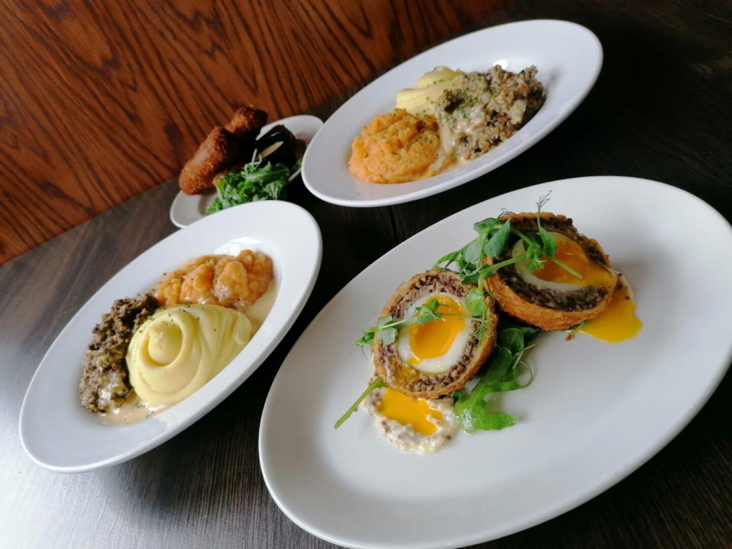 A table set with 2 plates of haggis, mashed potatoes and turnip and one of a open Scotch egg with salad garnish.