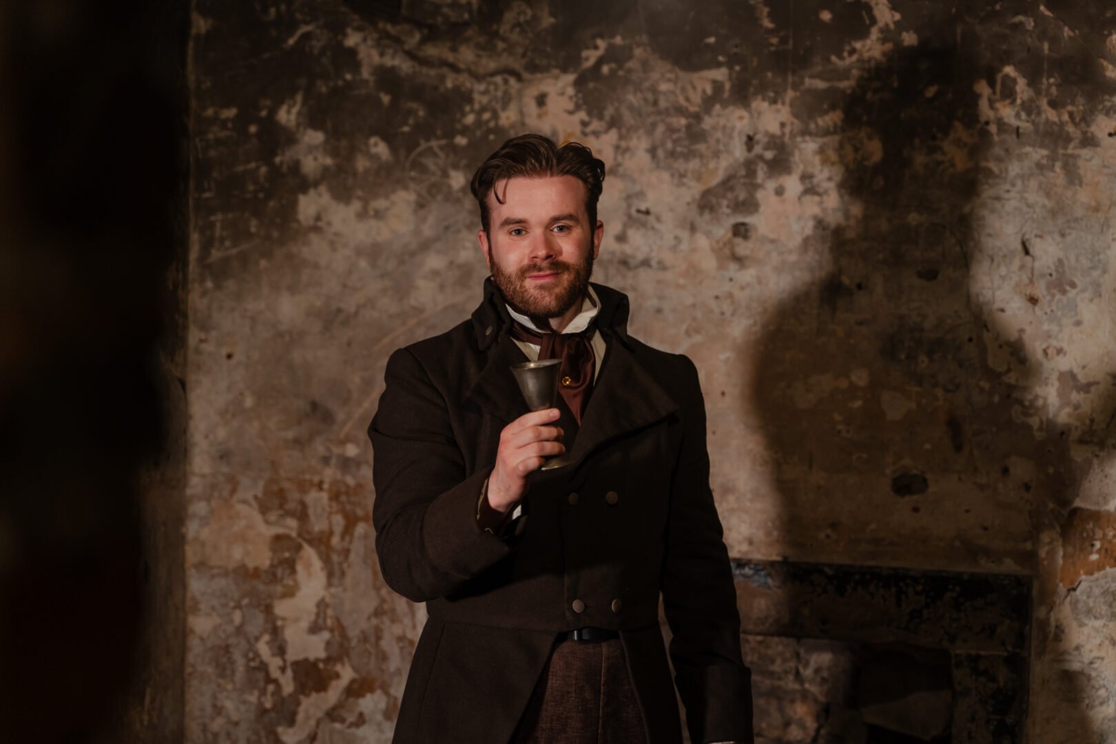 A man wearing an old fashioned long brown jacket, trousers and tie, holding a drinking cup.