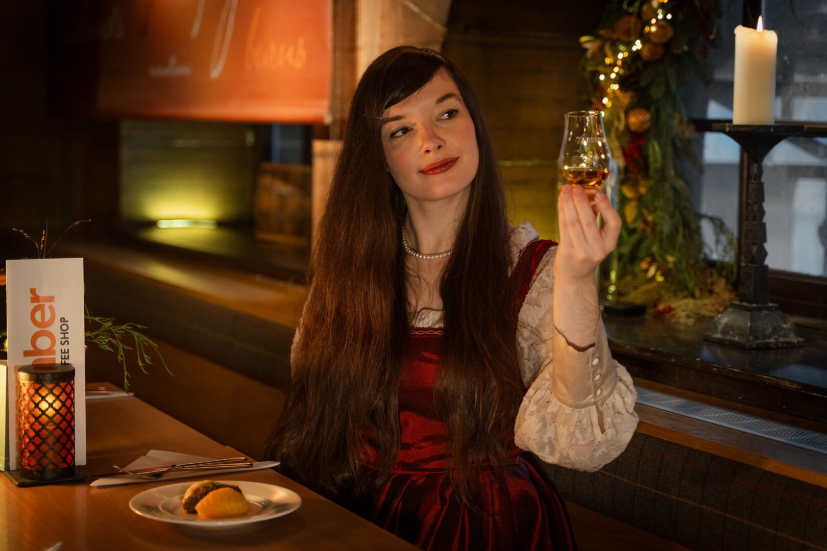 A women with long brown hair, red dress and cream coloured shirt sitting at a table holding aloft a glass filled with amber liquid.