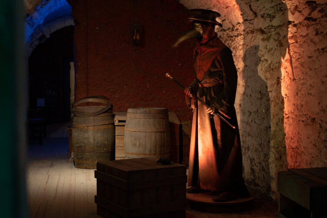 A tall figure wearing a mask with a pointed nose standing in a dimly lit room.