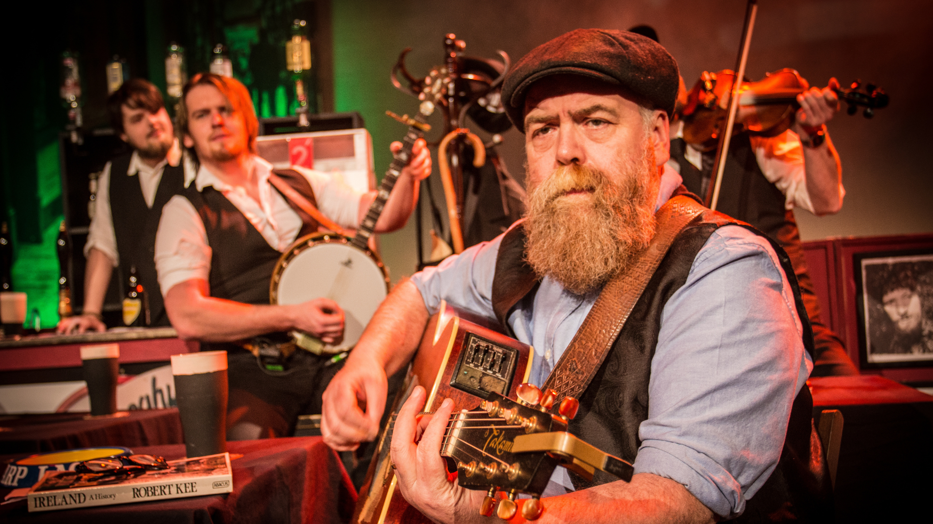 2 men, one with a bushy beard, in a pub. One is playing a guitar, and one a banjo.