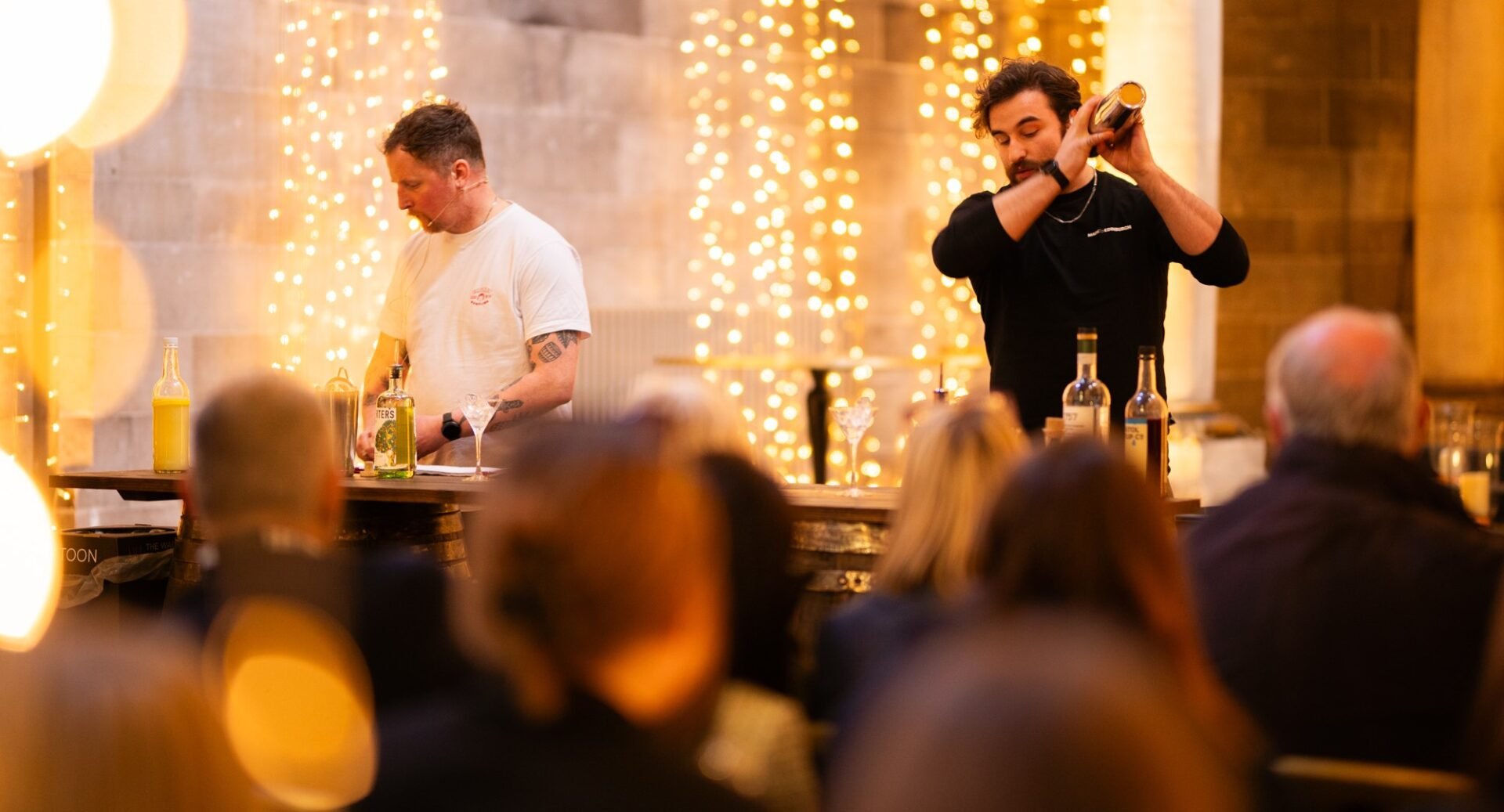 2 men standing at the front of a crowd of people, one of whom is shaking a cocktail shaker.