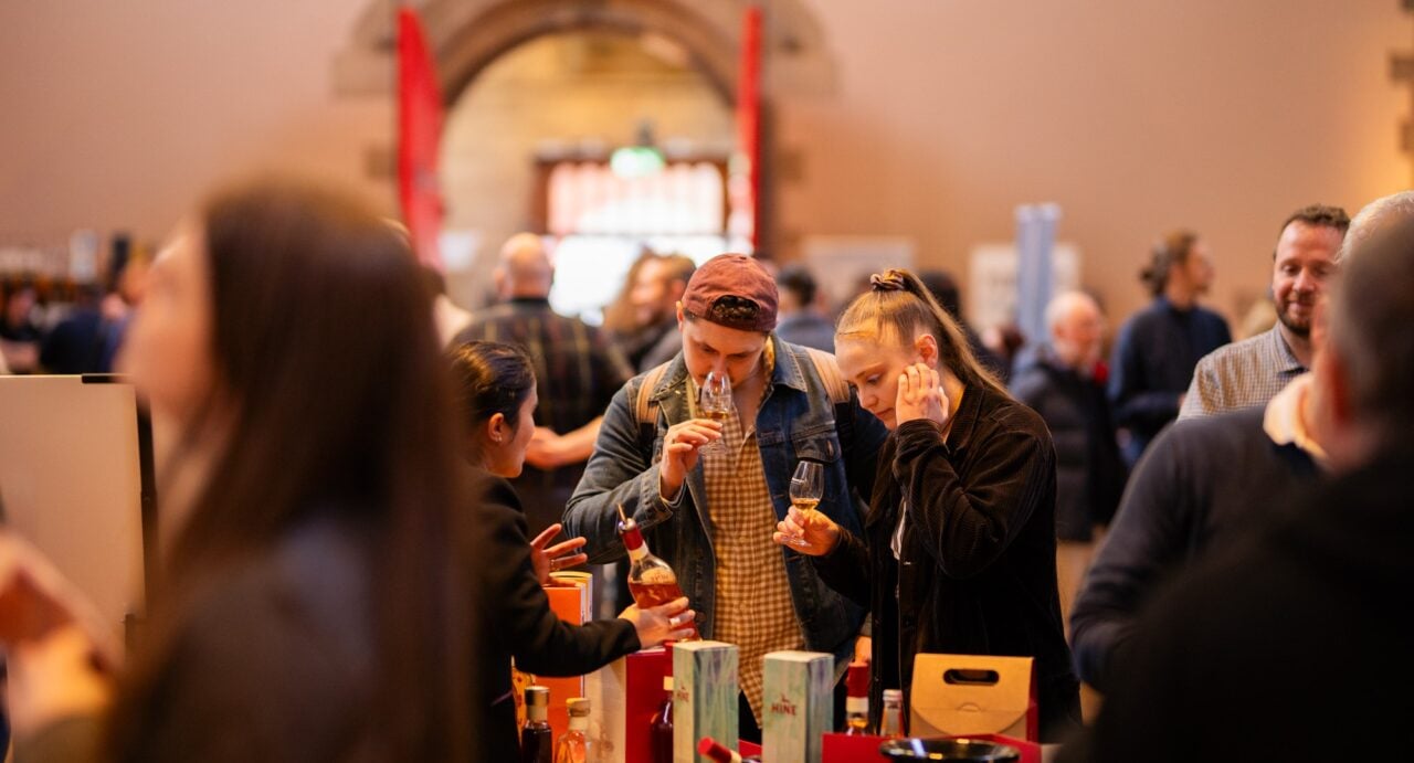 A crowd of people in a room. At the foreground are 2 people sniffing the contents of a glass, which is filled with an amber liquid.