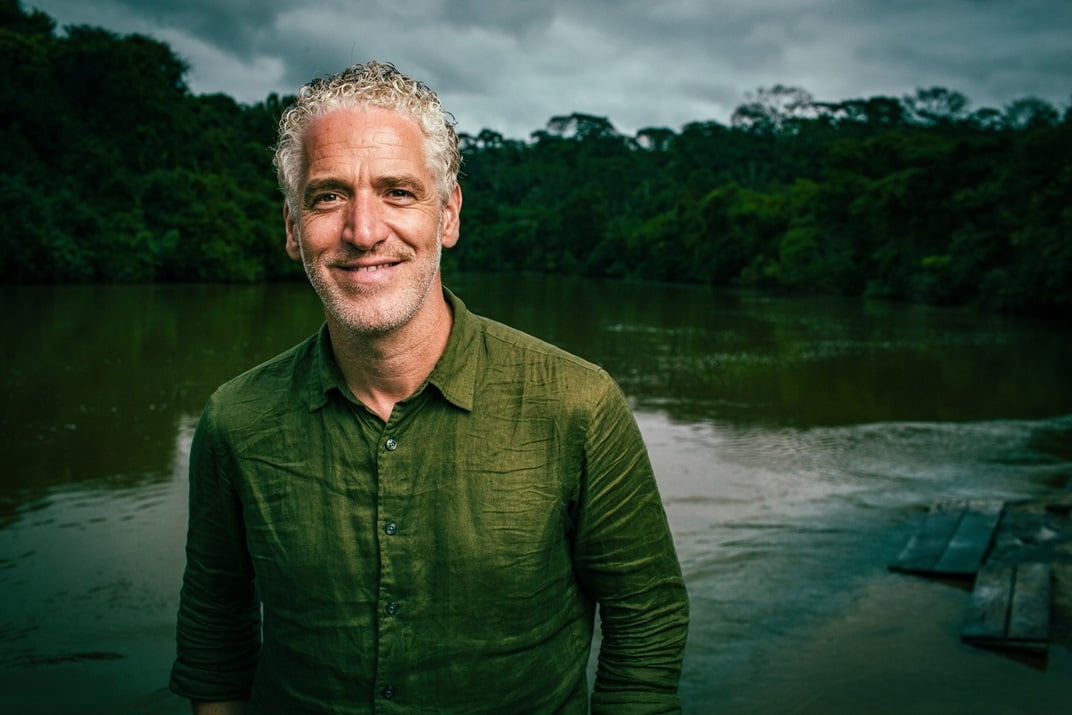 Image of man with grey curly hair, wearing a green shirt, standing against a river and tree background.