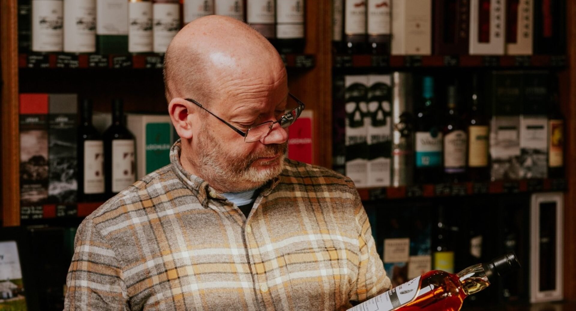 A man standing against a wall of bottled drinks, holding a bottle of whisky.