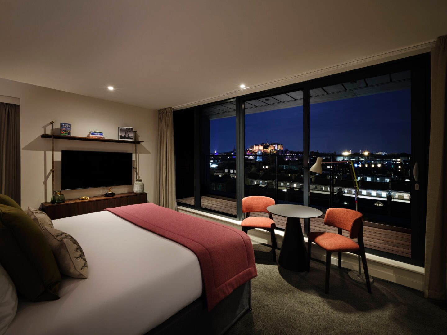 Night-time image of double bedroom with ceiling to floor window, looking out to city skyline.