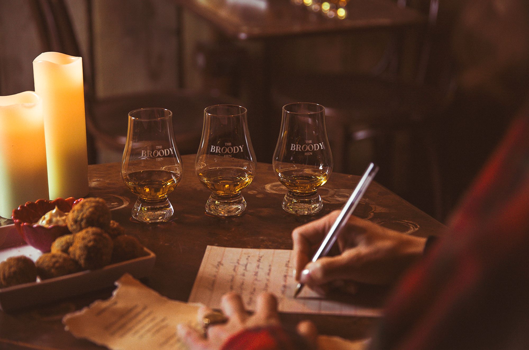 Person sitting at desk writing letter. On the table beside them are 3 glasses of whisky, a bowl of food and 2 lit candles.