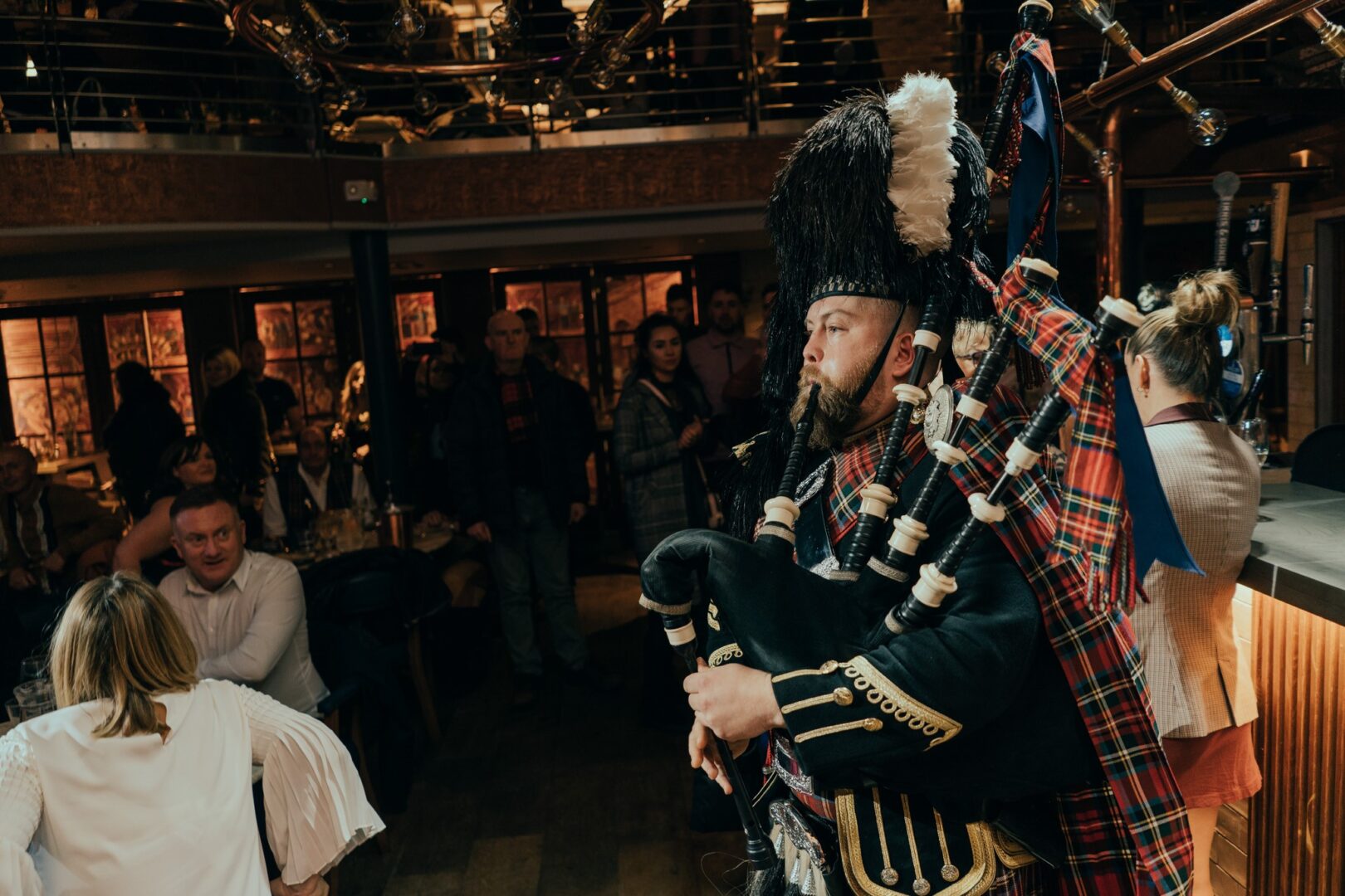 Piper in full Scottish dress playing pipes and entertaining guests in a bar.