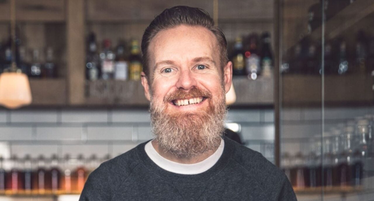 Head and shoulders image of man with brown/grey beard and brown hair, standing in front of a bar filled with shelves of bottles.