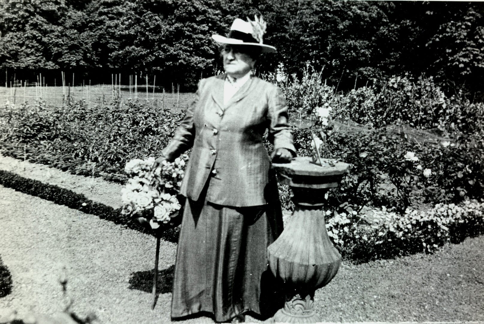 A black and white photo of Mrs Reid with cane and straw hat wearing a jacket and long skirt and standing beside a stone sundial